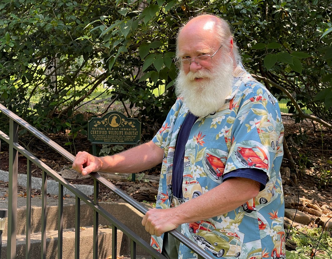 Ken Barker is pictured, an older, white, balding man with white hair and beard. He is standing outside with greenery in background wearing a short-sleeved patterned shirt.