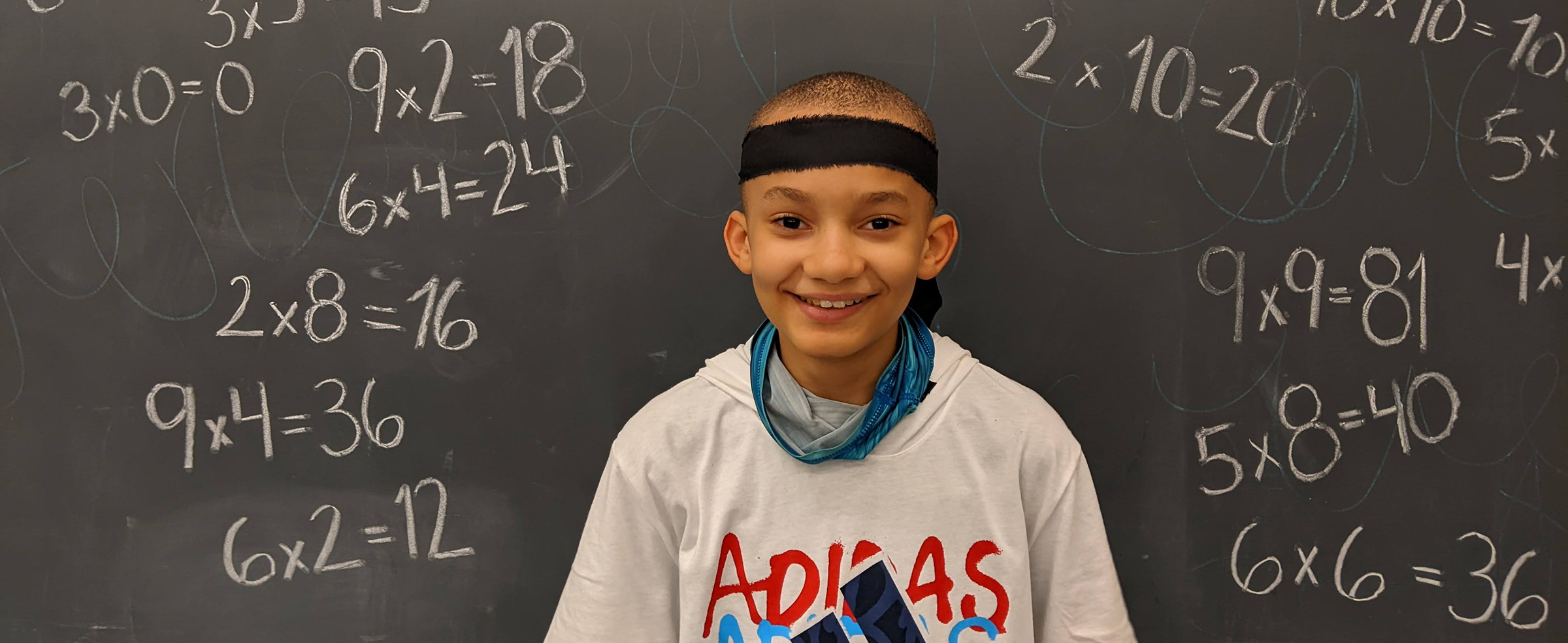 Young boy wearing a headband and wearing an adidas shirt stands smiling in front of a chalkboard covered in math problems.
