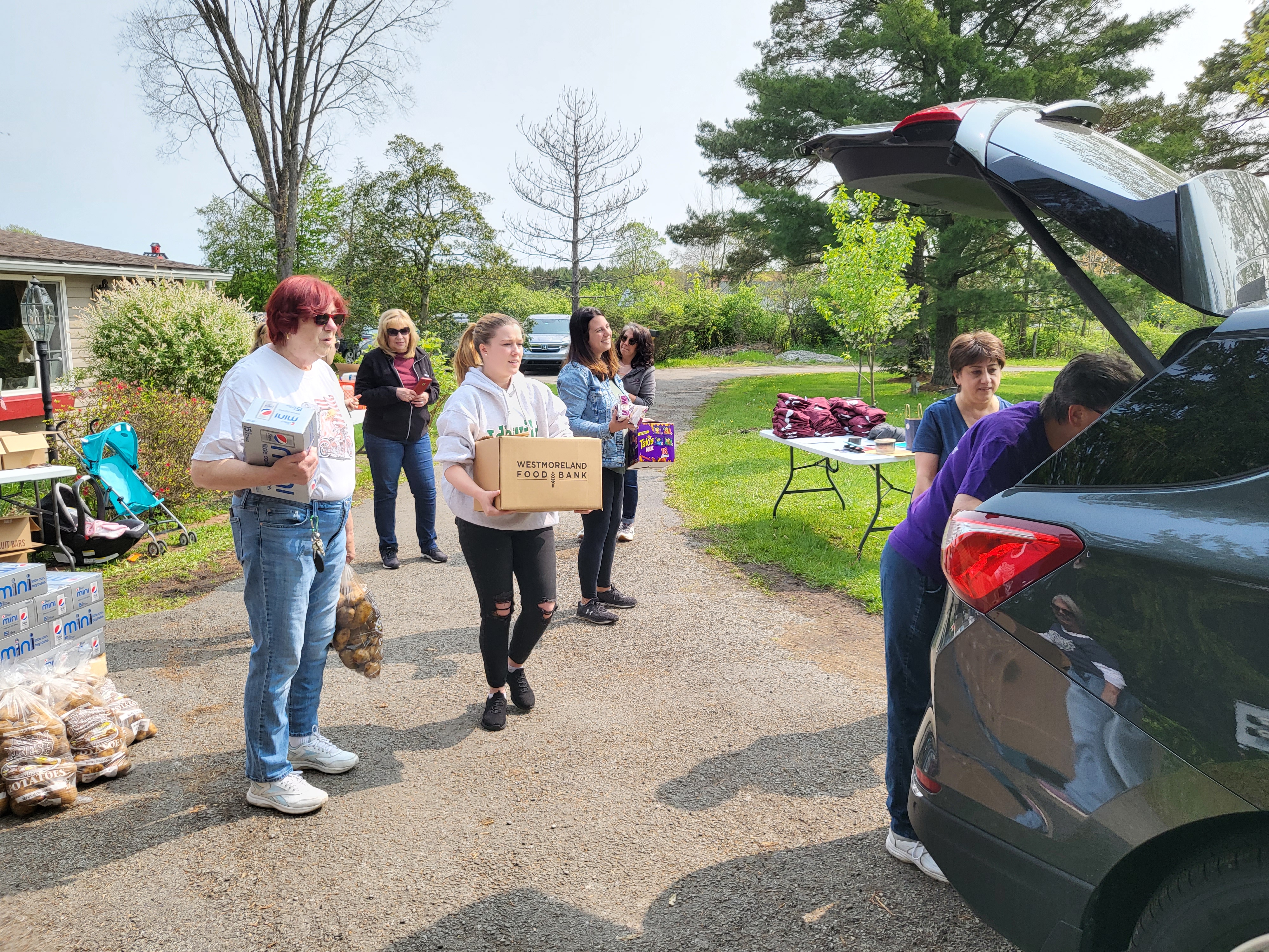 A group of Westmoreland Food Bank volunteers are gathered around a car with an open trunk. One volunteer in the middle of the photo is carrying a box of food to the car.