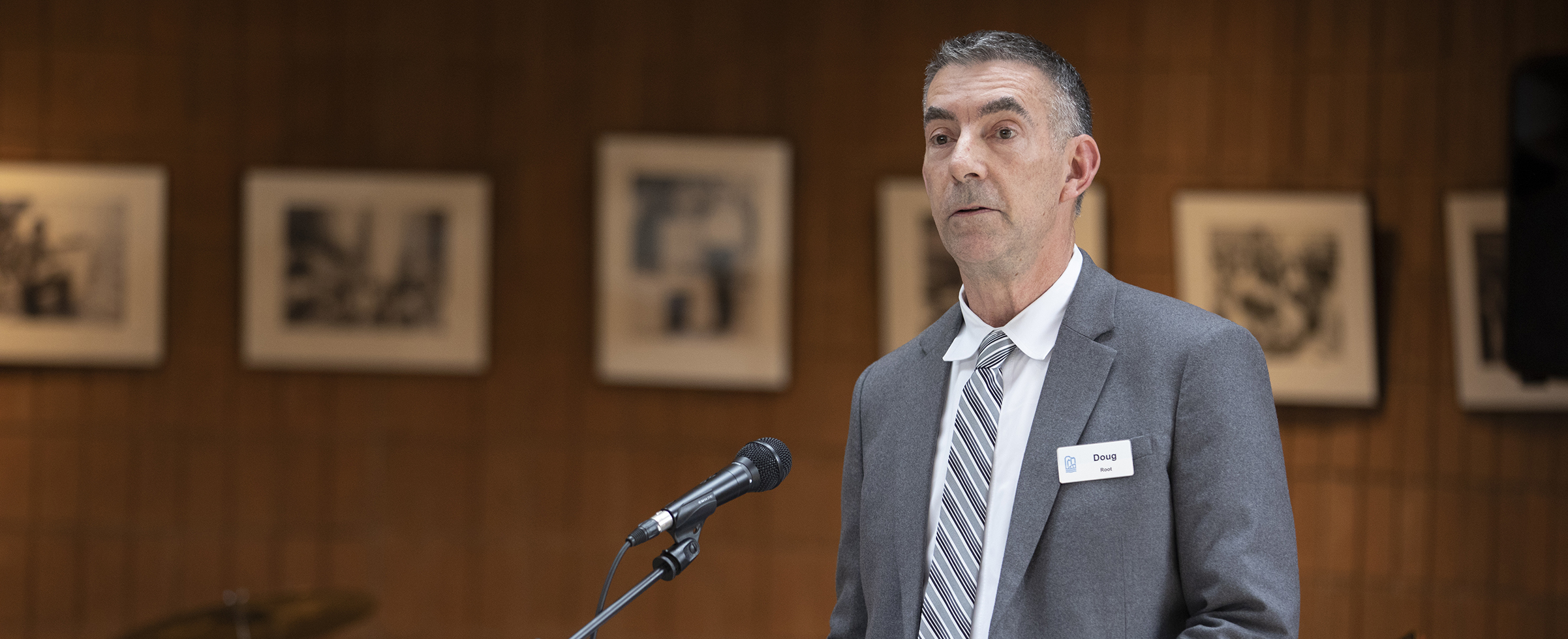 Man with short dark hair speaking at a podium, wearing blue jacket, white shirt and tie.