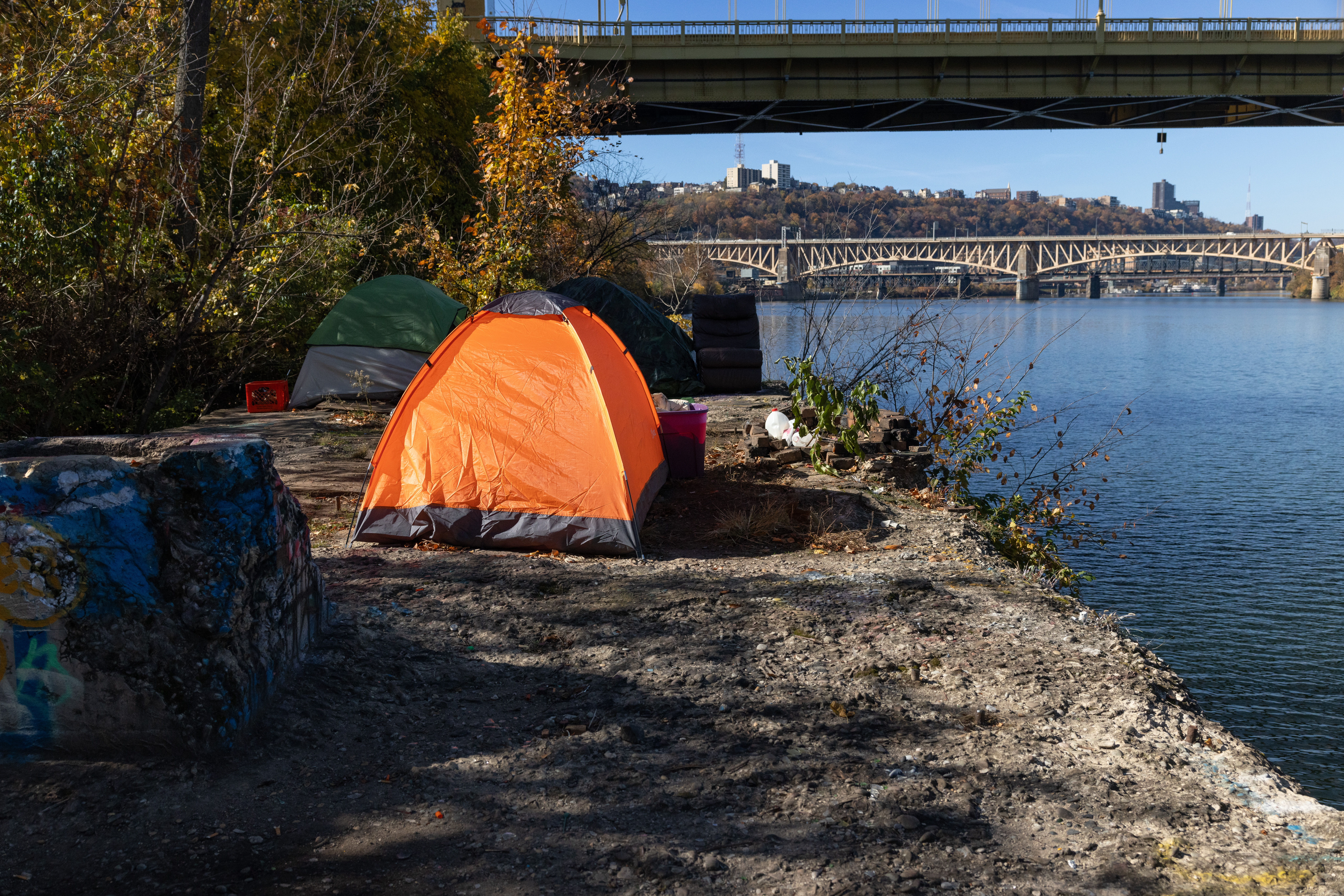 A bright orange tent sits by the riverbank with a sleeping bag and other personal items nearby