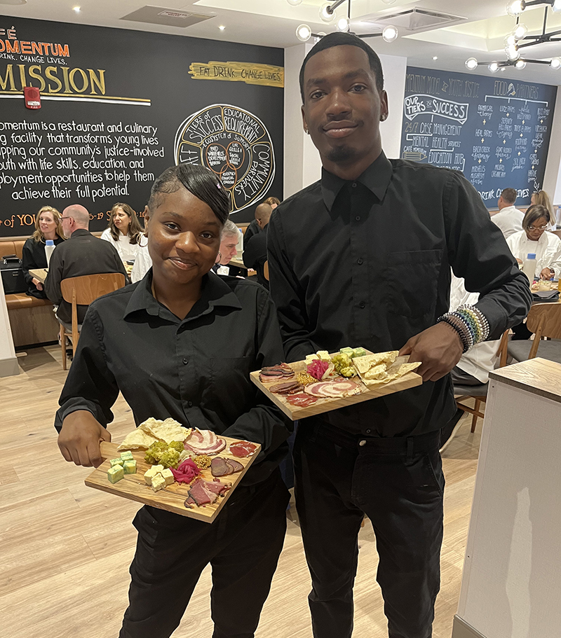 Young woman and man standing and holding platters of food they are serving at Cafe Momentum.