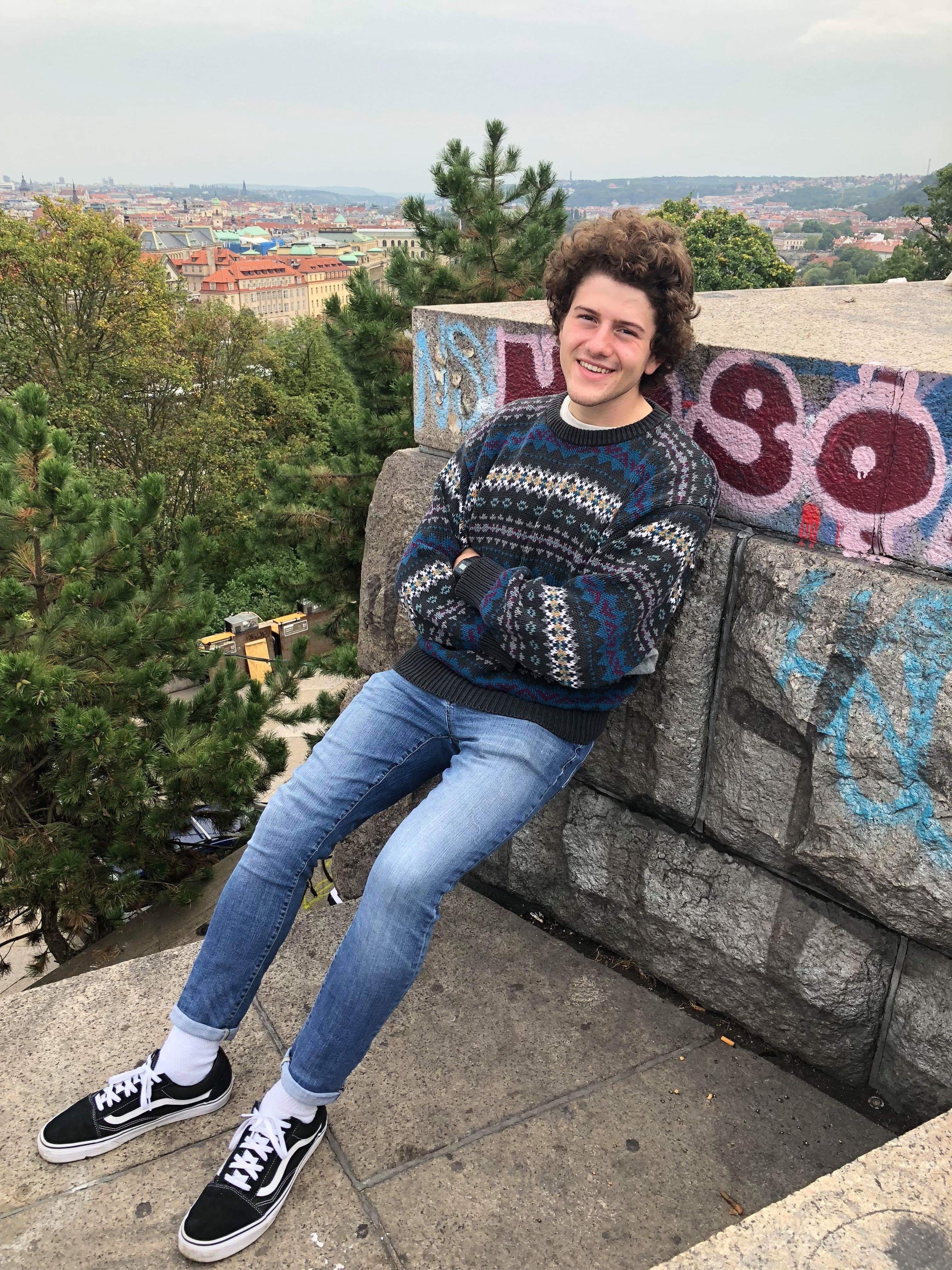 Christian Cochran, standing outside, leaning up against a rock wall, wearing blue jeans and sneakers.