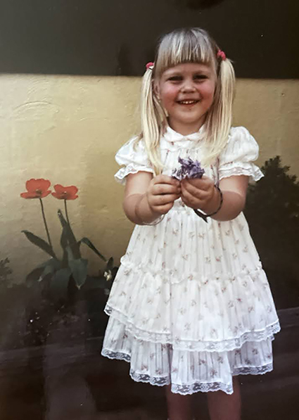 Young, blonde girl with hair in pony tails, holding a flower and wearing a white dress. 
