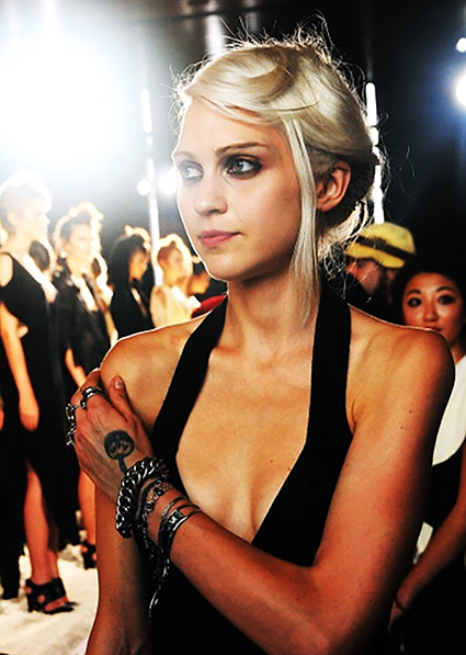 Young woman with blonde hair, wearing black dress and chain-style bracelets. 