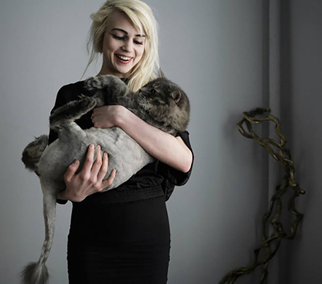 Young woman with blonde hair, holding a cat and smiling at it. 