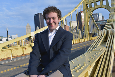 Young, smiling teenage male dressed in a blue sport coat, Richie Hrivnak, sitting outside on top of a yellow bridge support with Pittsburgh skyline in background.