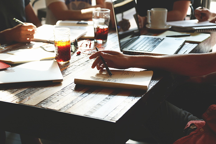Showing hands and arms of two people working on a desk and writing in notebooks and working on a laptop.