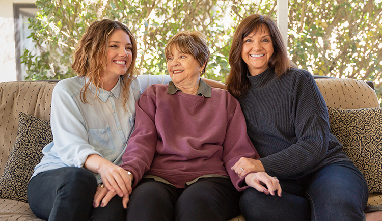 Daughters sit on a couch with their mother iin the middle. All three are smiling.