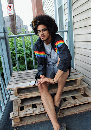 Young man sitting on steps outside a house wearing shorts and a jacket with rainbow stripes.