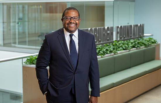 William Generett, Jr. standing in front of a Pittsburgh Foundation sign. Man with short, dark hair and wearing glasses and a dark blue suit with blue tie and white shirt. 